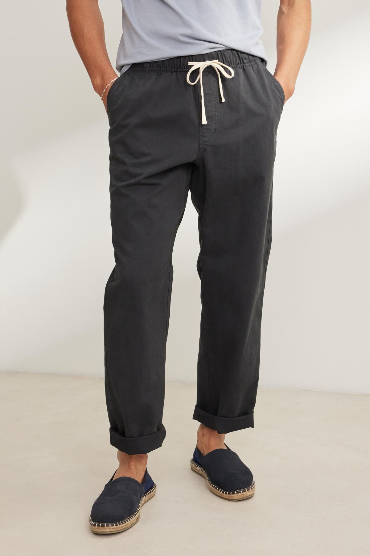   A person is wearing Velvet by Graham & Spencer's BRANSON PANT in a dark, straight-leg design made from cotton sanded twill, along with black slip-on shoes featuring white soles, standing on a light floor. 