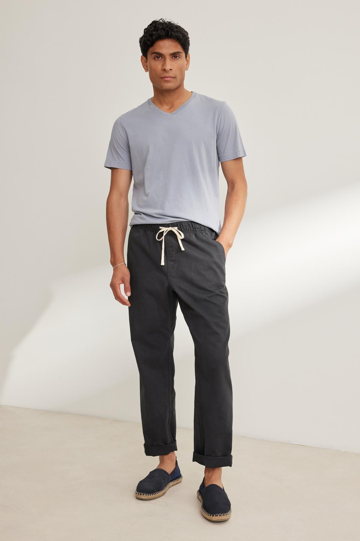 A person stands against a plain background dressed in light gray T-shirt, Velvet by Graham & Spencer's dark BRANSON PANT with an elastic waist and drawstring, paired with casual shoes.-38643232473281