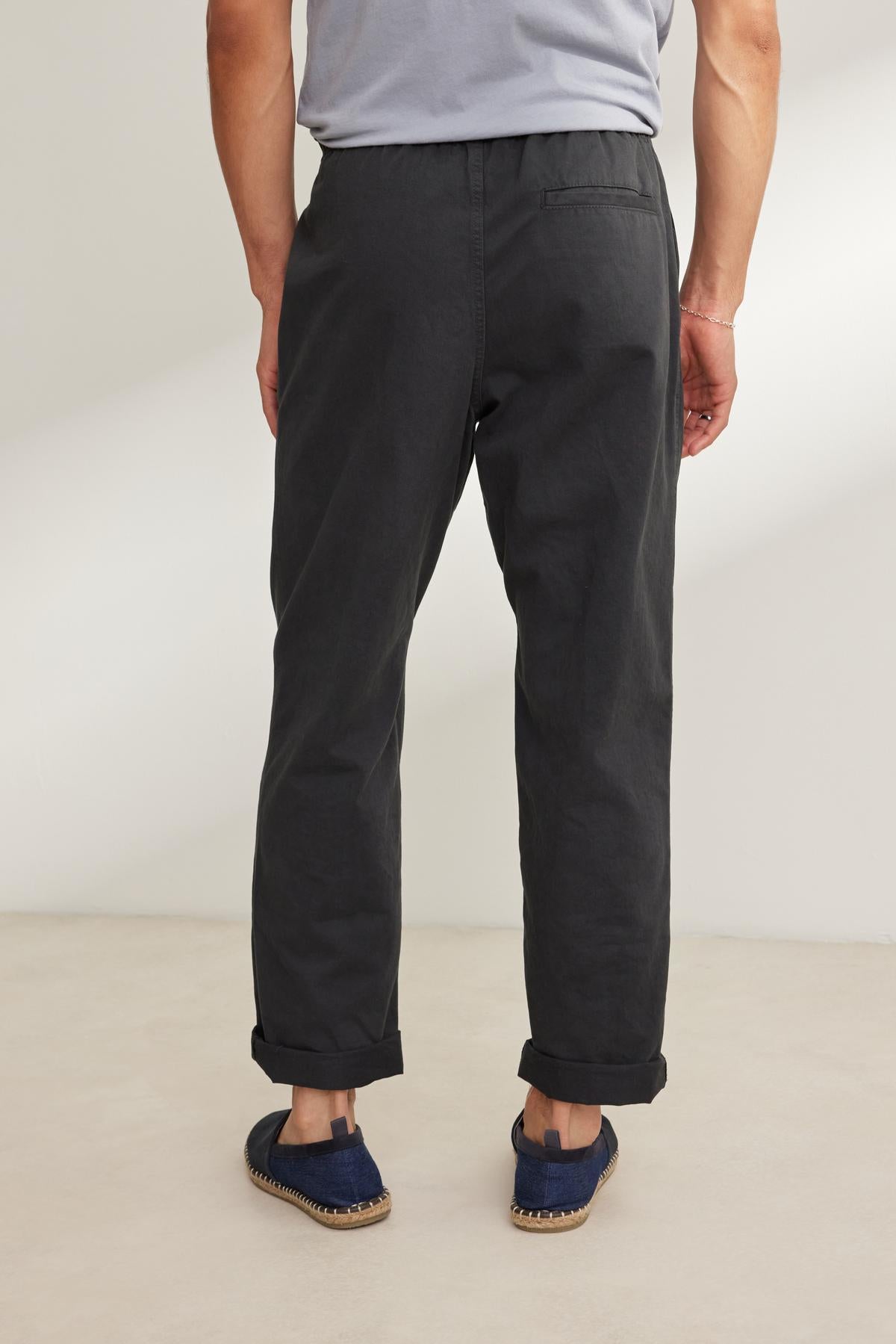 A person in a gray T-shirt and blue shoes stands on a light-colored floor with their back to the camera, wearing Velvet by Graham & Spencer's BRANSON PANT. These dark pants feature an elastic drawstring waist and straight leg design, effortlessly blending comfort with style.-38643232342209