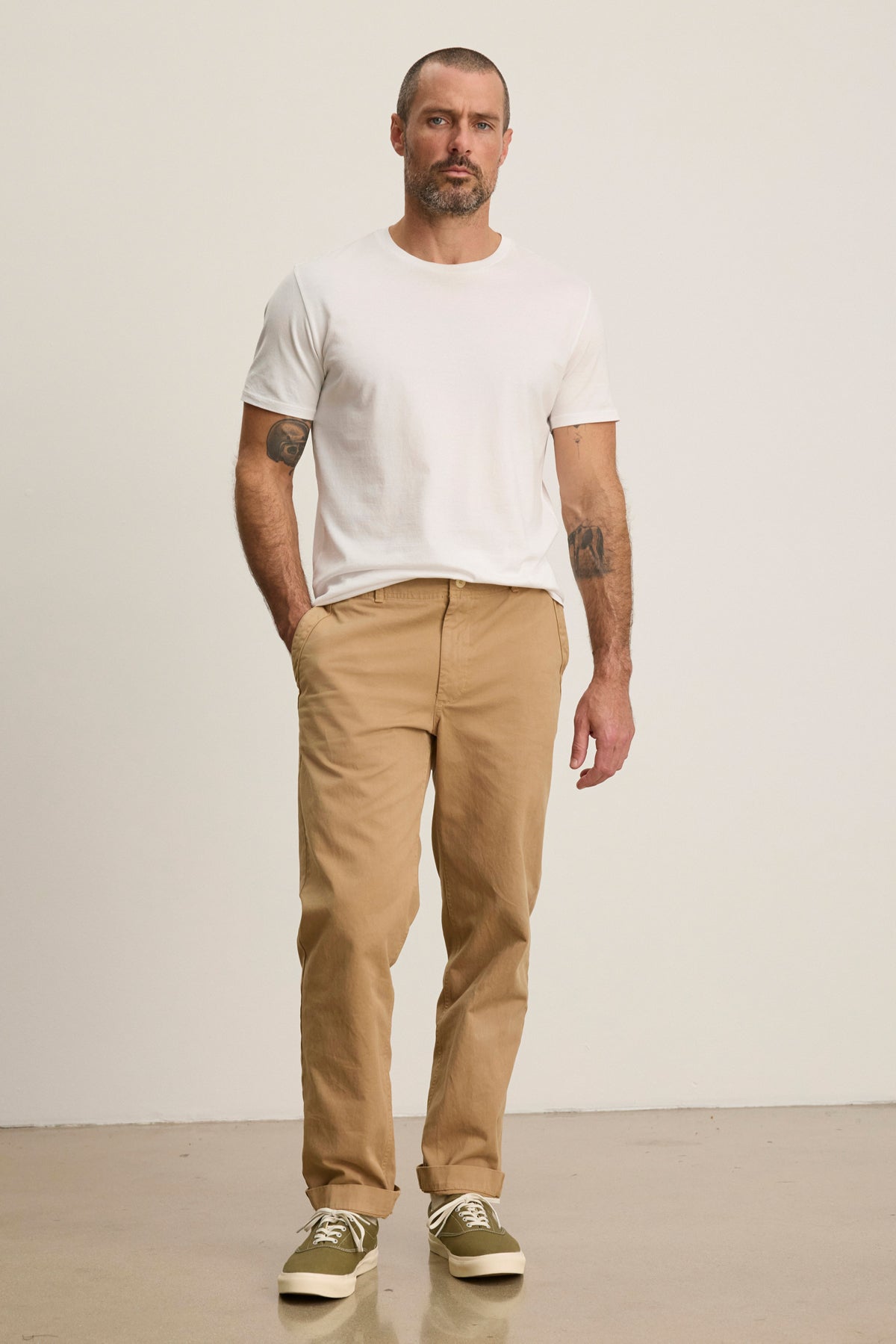 A man stands against a plain background wearing the LENNOX PANT by Velvet by Graham & Spencer—relaxed fit tan cotton twill pants—paired with a white t-shirt and beige sneakers, one hand in his pocket for an effortlessly stylish everyday look.-39446033465537