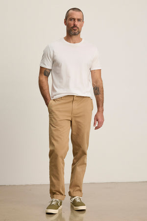 A man stands against a plain background wearing the LENNOX PANT by Velvet by Graham & Spencer—relaxed fit tan cotton twill pants—paired with a white t-shirt and beige sneakers, one hand in his pocket for an effortlessly stylish everyday look.