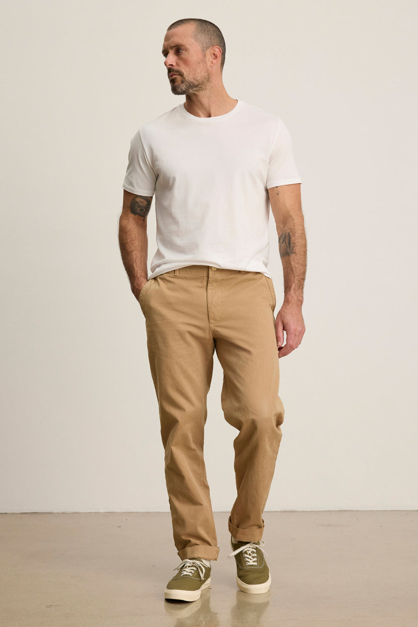 A man stands against a plain background wearing the LENNOX PANT by Velvet by Graham & Spencer in tan cotton twill, styled with a relaxed white tee and olive sneakers—an ideal look for effortless, everyday wear.