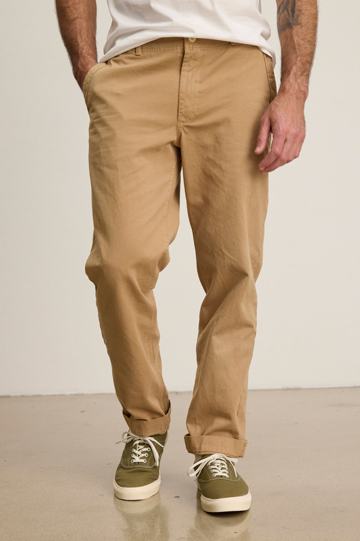 A person wears Velvet by Graham & Spencer LENNOX PANT in relaxed fit beige cotton twill, paired with a white shirt and green sneakers, standing with hands in pockets on a light floor.-39446033531073