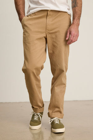 A person wears Velvet by Graham & Spencer LENNOX PANT in relaxed fit beige cotton twill, paired with a white shirt and green sneakers, standing with hands in pockets on a light floor.