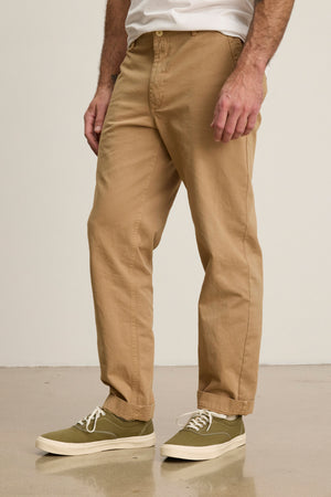 Indoors on a polished floor, a person wears Velvet by Graham & Spencer's LENNOX PANT—relaxed fit tan cotton twill pants—paired with olive green sneakers with white soles, ideal for everyday style.