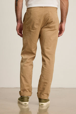 A person wearing the LENNOX PANT by Velvet by Graham & Spencer in relaxed fit tan cotton twill, paired with a white shirt and green sneakers, stands on a light-colored floor facing away—ideal for everyday wear.