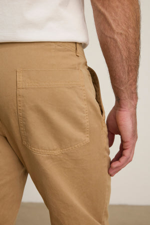 A close-up of a right hand near the back pocket of the LENNOX PANT by Velvet by Graham & Spencer in tan, paired with a white shirt—an ideal choice for everyday wear.