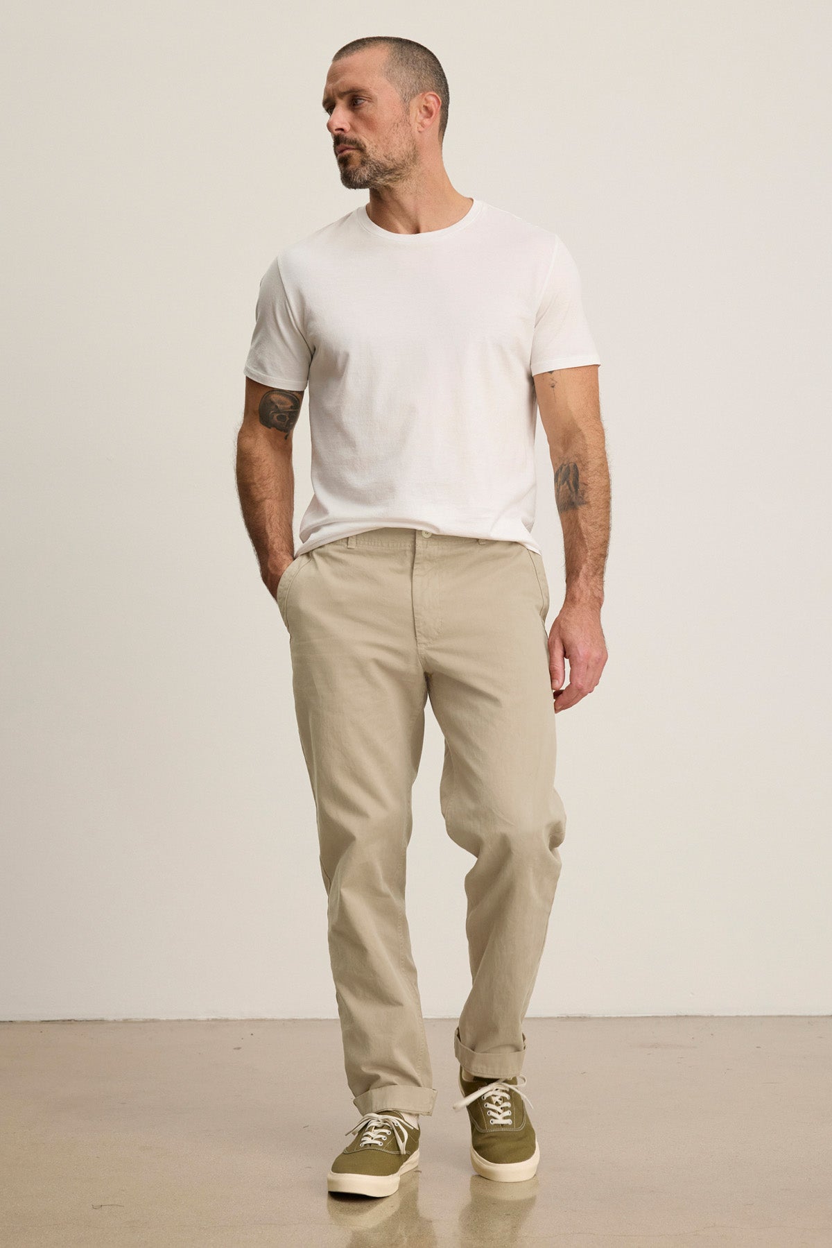 A man stands against a plain background in a white t-shirt, Velvet by Graham & Spencer LENNOX PANT, and olive green sneakers with hands in pockets—an ideal look for everyday wear.-40850767282369