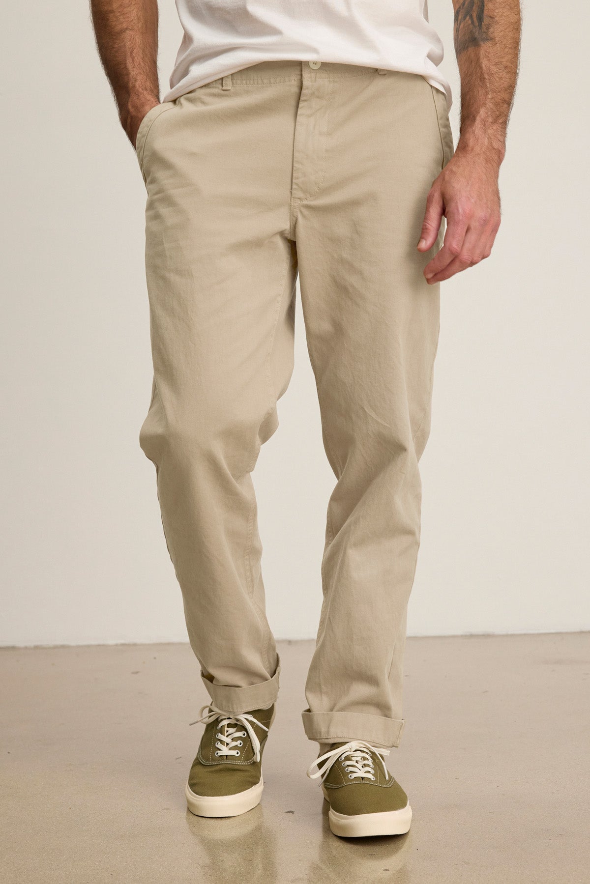 A man wearing the LENNOX PANT by Velvet by Graham & Spencer and green sneakers with white laces stands with one hand in his pocket against a plain background.-40850767249601
