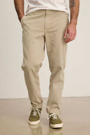 A man wearing the LENNOX PANT by Velvet by Graham & Spencer and green sneakers with white laces stands with one hand in his pocket against a plain background.