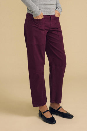 A person wearing a light gray sweater and the BRYLIE PANT by Velvet by Graham & Spencer—burgundy cotton twill trousers featuring a barrel leg silhouette—paired with black Mary Jane shoes, stands with hands in pockets against a beige background.