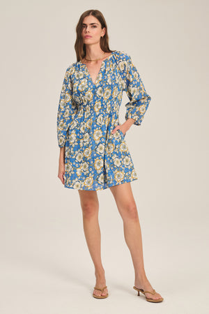 A woman wears the SANDRINE DRESS by Velvet by Graham & Spencer, featuring a blue floral pattern, long sleeves, V-neckline, and short hemline. She pairs it with tan sandals.