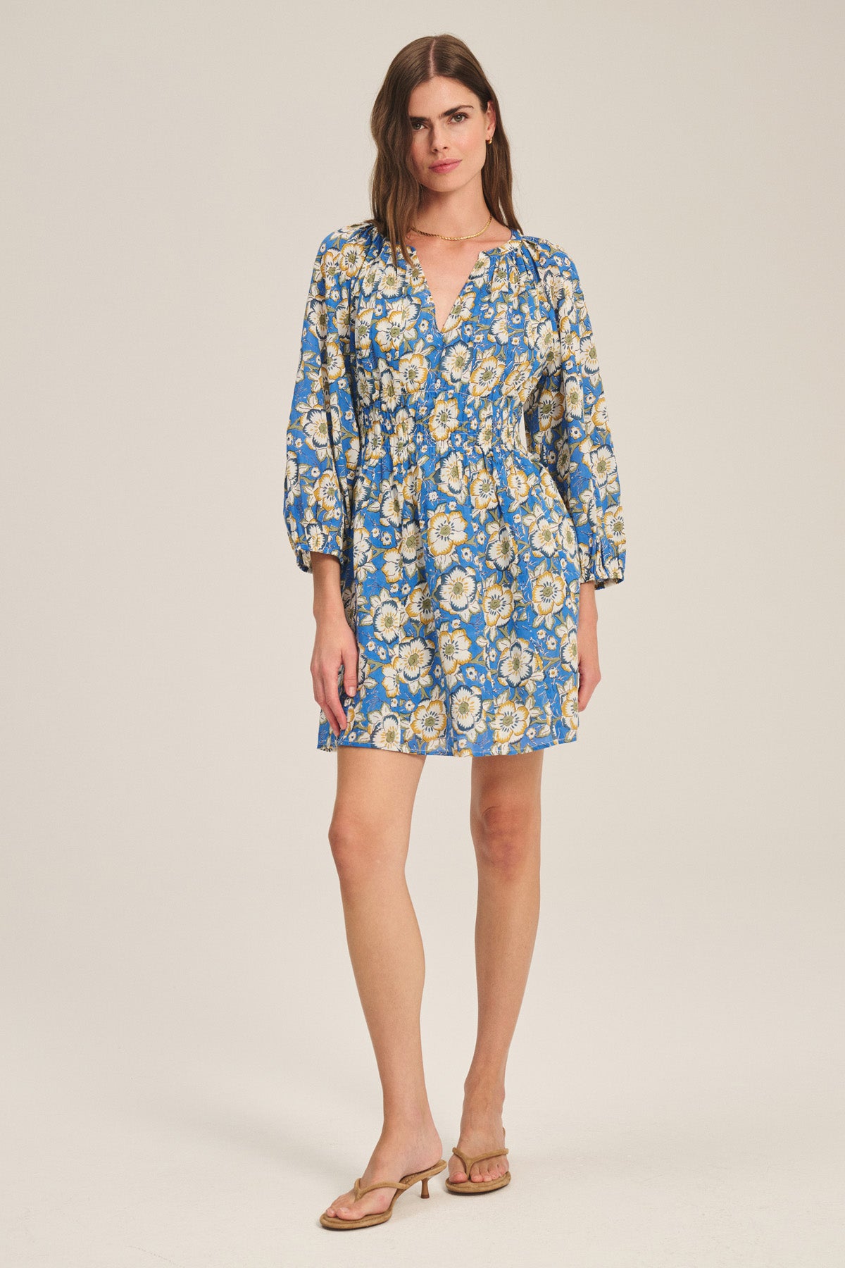 A woman wears the SANDRINE DRESS by Velvet by Graham & Spencer, a blue floral long-sleeve mini dress with a V-neckline, paired with tan heeled sandals, standing against a plain light background.