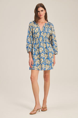 A woman wears the SANDRINE DRESS by Velvet by Graham & Spencer, a blue floral long-sleeve mini dress with a V-neckline, paired with tan heeled sandals, standing against a plain light background.