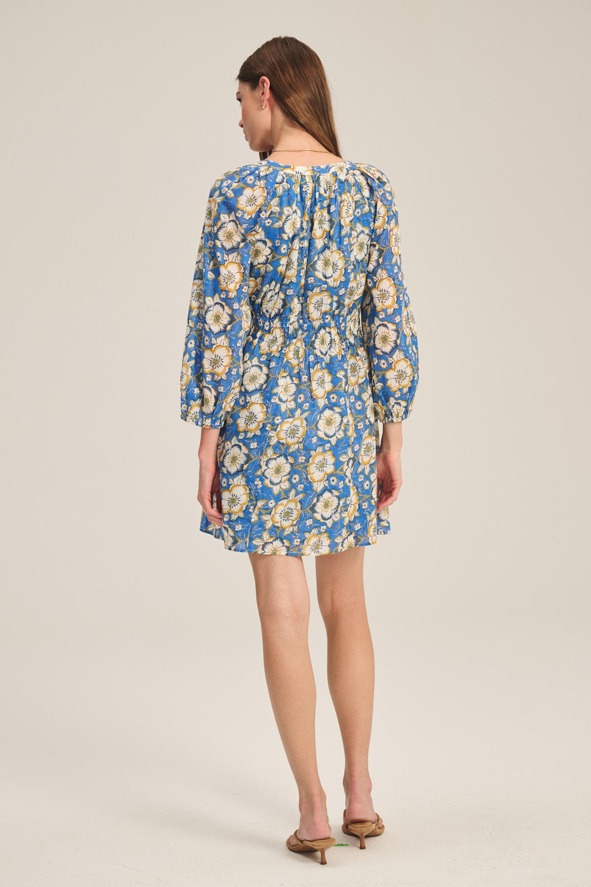 A woman faces away, wearing the SANDRINE DRESS by Velvet by Graham & Spencer—a blue floral long-sleeve dress—paired with brown heeled sandals against a plain background.