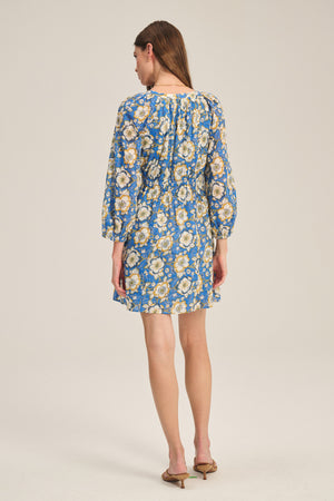 A woman faces away, wearing the SANDRINE DRESS by Velvet by Graham & Spencer—a blue floral long-sleeve dress—paired with brown heeled sandals against a plain background.