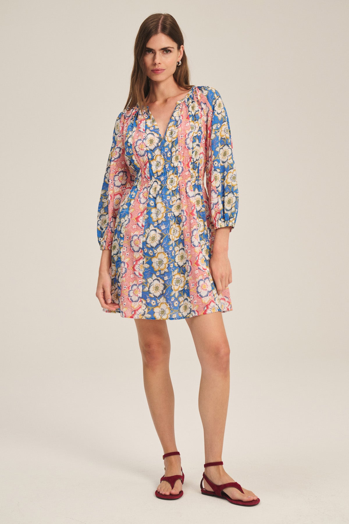 A woman wears the Velvet by Graham & Spencer SANDRINE DRESS—a long-sleeve, floral mini dress in pink, blue, and yellow—paired with red flat sandals, standing against a plain light background.-41436017950913