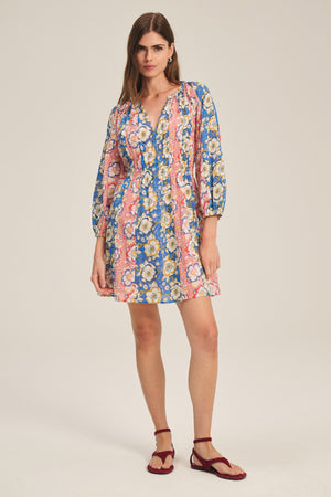 A woman wears the Velvet by Graham & Spencer SANDRINE DRESS—a long-sleeve, floral mini dress in pink, blue, and yellow—paired with red flat sandals, standing against a plain light background.