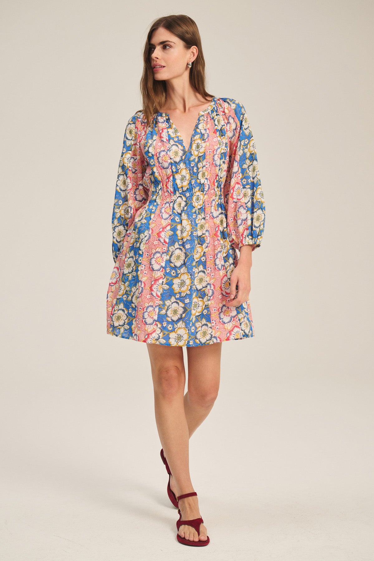 A woman wearing the SANDRINE DRESS by Velvet by Graham & Spencer—a long-sleeve, floral print mini dress in pink, blue, and yellow with a V-neckline—stands with one leg slightly bent.
