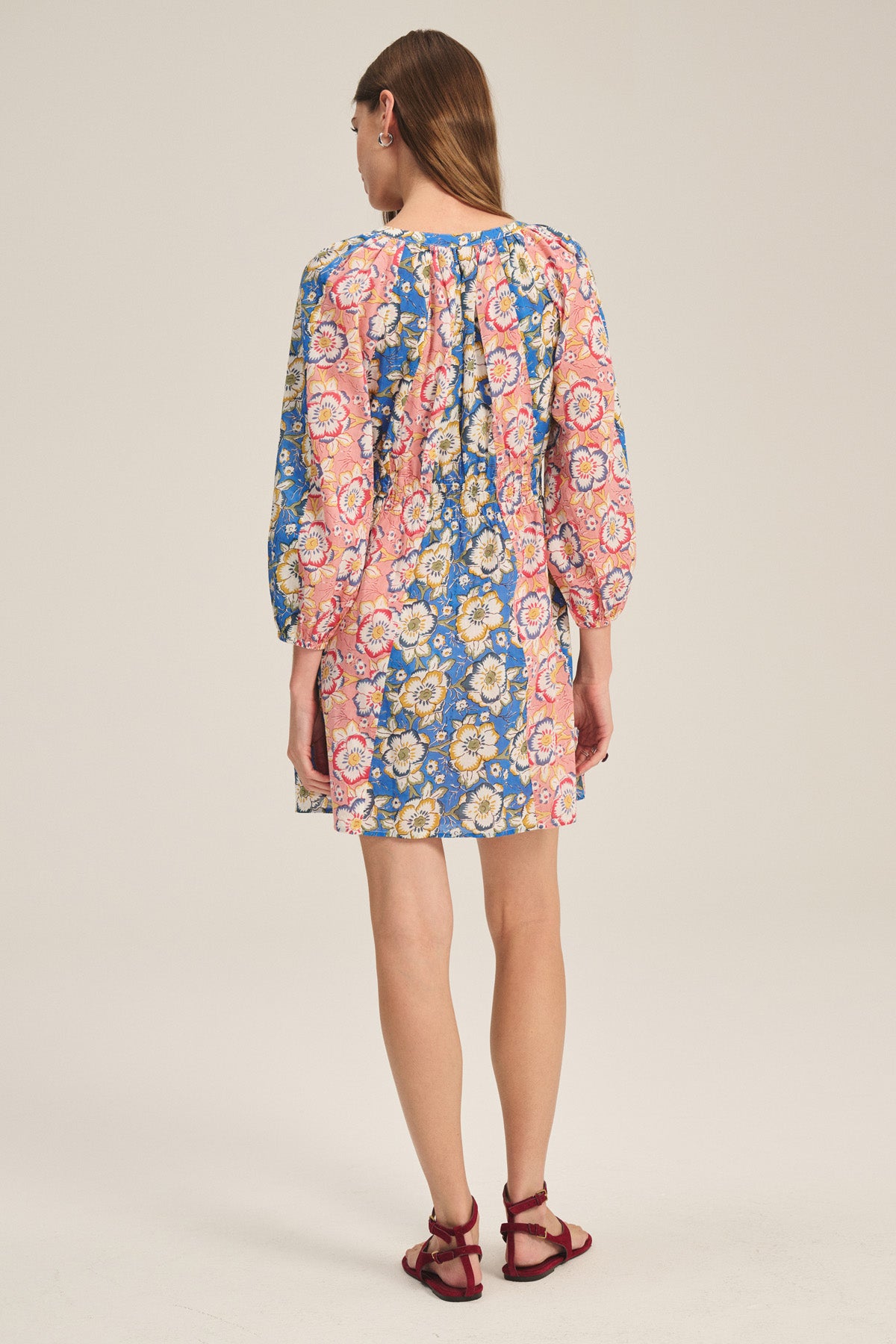 A woman faces away, wearing the SANDRINE DRESS by Velvet by Graham & Spencer—a colorful, long-sleeve floral dress with a round neckline—paired with red sandals against a plain background.-41436015984833