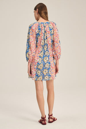 A woman faces away, wearing the SANDRINE DRESS by Velvet by Graham & Spencer—a colorful, long-sleeve floral dress with a round neckline—paired with red sandals against a plain background.