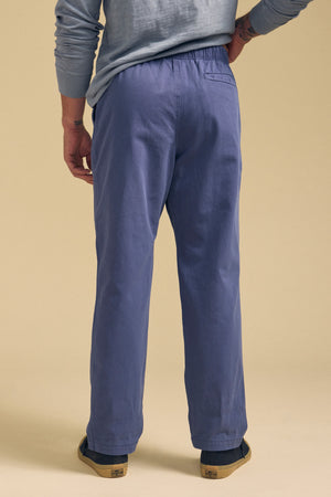 A person stands facing away, wearing Velvet by Graham & Spencer's BRANSON PANT with a straight leg and elastic drawstring waist, paired with a light blue long-sleeve shirt and slip-on shoes against a plain beige background.