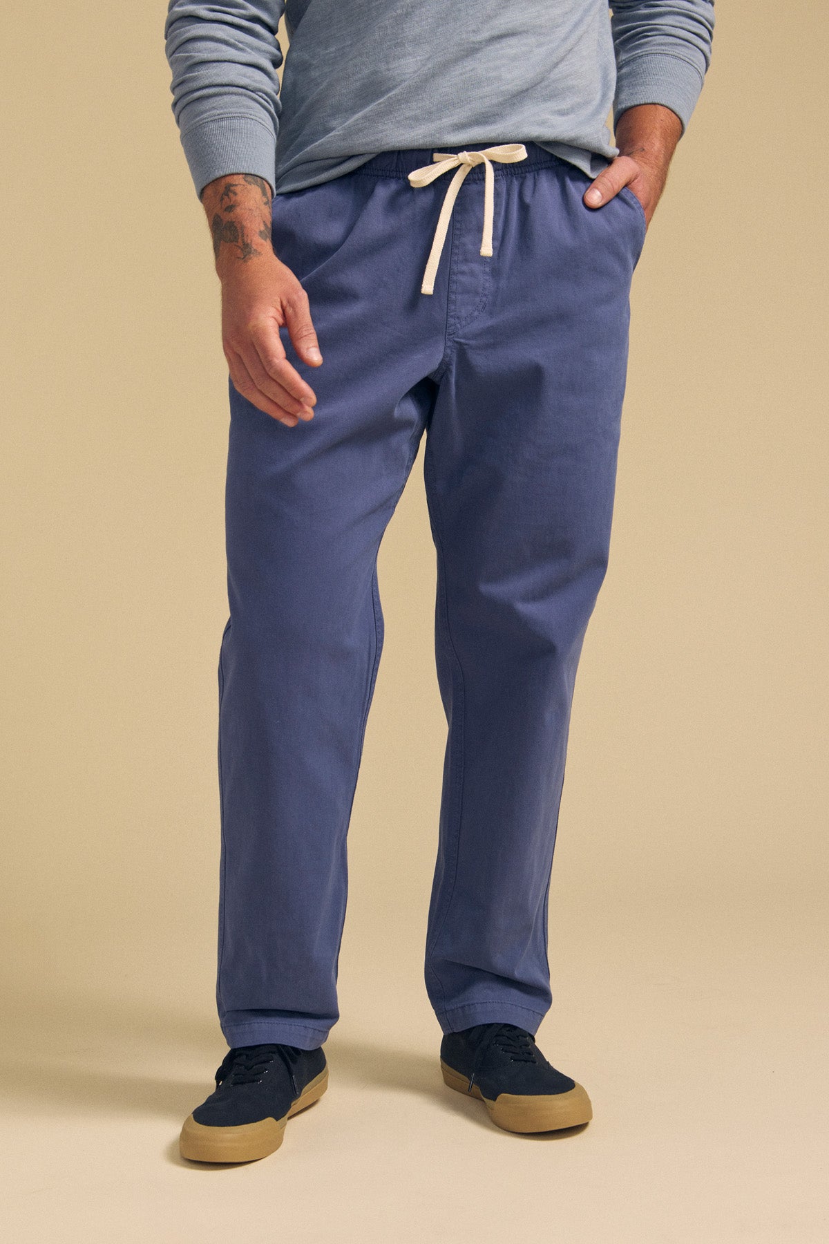 A man wearing Velvet by Graham & Spencer's BRANSON PANT in blue, a light blue long-sleeve shirt, and navy sneakers stands with one hand in his pocket against a beige background.