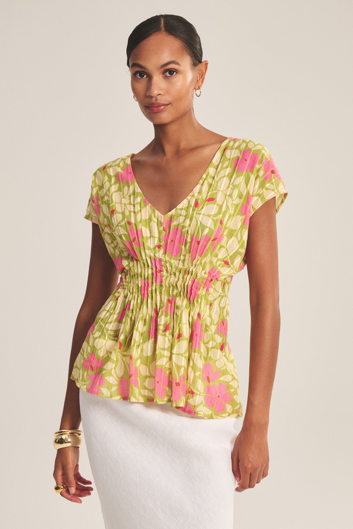 A woman wears the SARAH TOP by Velvet by Graham & Spencer—a short-sleeve, V-neck green and pink floral cotton blouse with pleated waist—paired with a white skirt and gold accessories against a plain background.