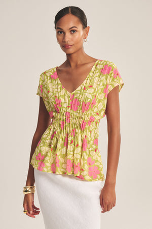 A woman wears the SARAH TOP by Velvet by Graham & Spencer—a short-sleeve, V-neck green and pink floral cotton blouse with pleated waist—paired with a white skirt and gold accessories against a plain background.