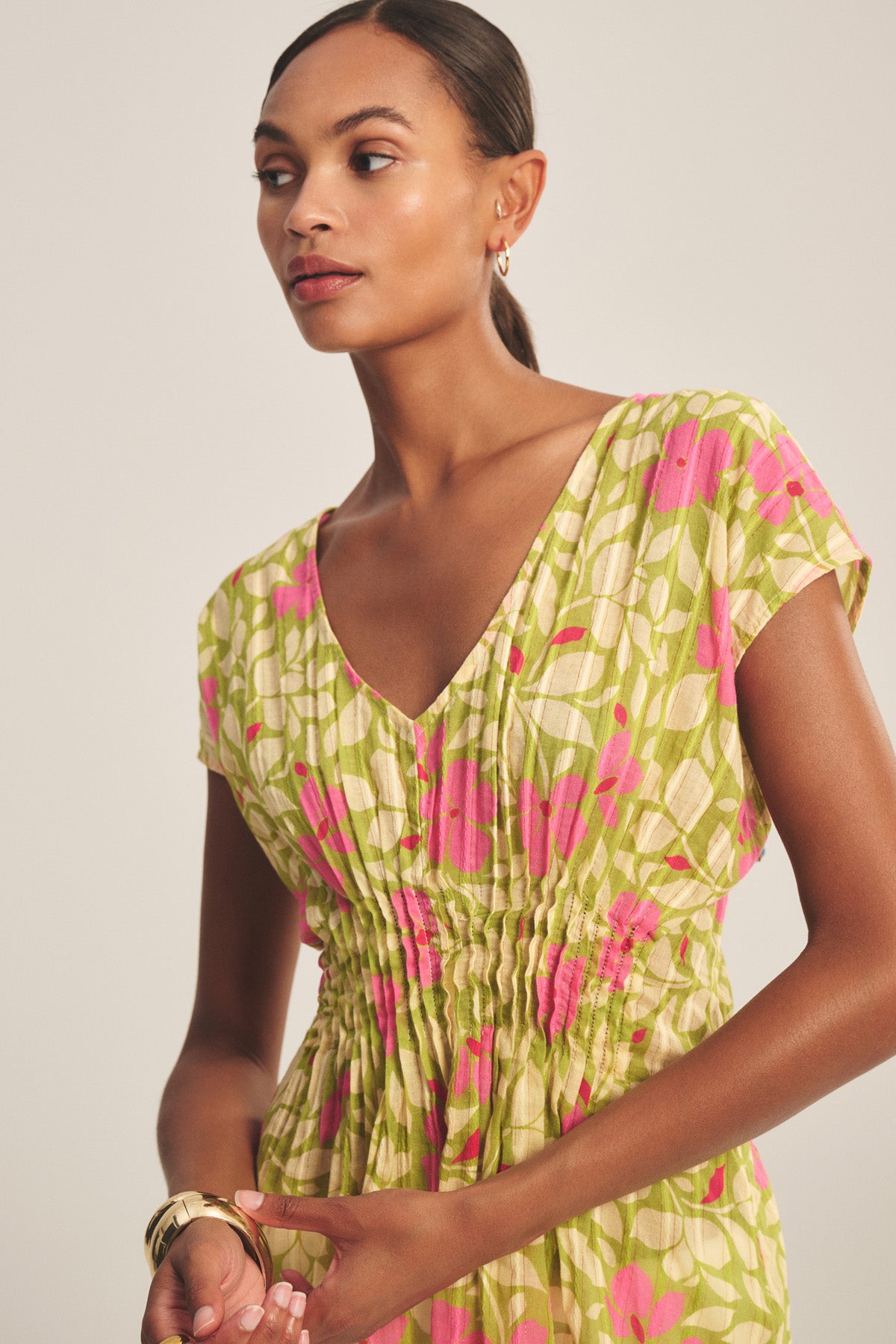 A woman wears the Velvet by Graham & Spencer SARAH TOP with a V-neck, short sleeves, pink and green floral pattern, and pleated waist, standing against a neutral background.