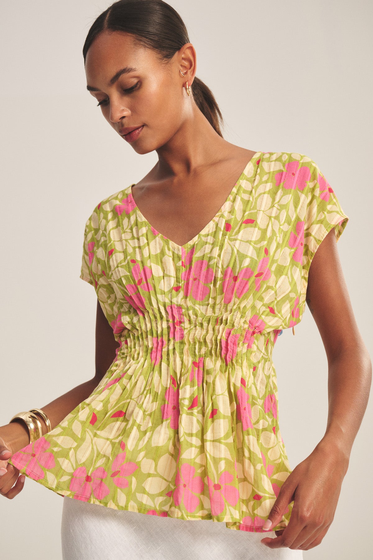 A woman wears the SARAH TOP by Velvet by Graham & Spencer—light green and pink floral cotton with short sleeves, V-neck, and pleated waist—paired with white bottoms and gold hoop earrings.-41128319975617