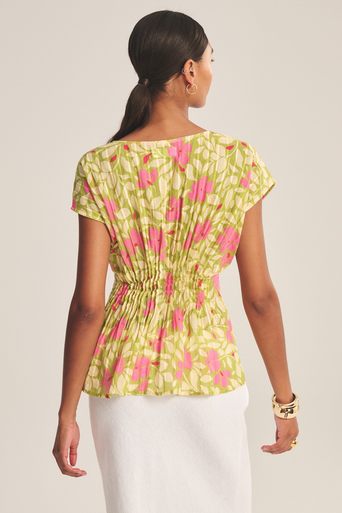 A woman faces away, wearing the Velvet by Graham & Spencer SARAH TOP—a green and pink patterned cotton piece with a gathered, pleated waist—paired with a white skirt, gold hoop earrings, and gold bracelets.