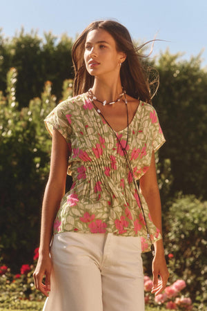 A woman stands outdoors in a sunny garden wearing the SARAH TOP by Velvet by Graham & Spencer—a short-sleeved, pink and green patterned cotton top with a pleated waist—paired with white pants.