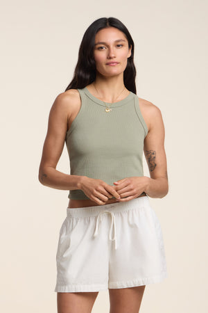 A woman wears the SARI TANK TOP by Velvet by Graham & Spencer, a light green ribbed cotton cropped tank, paired with white drawstring shorts against a plain background.