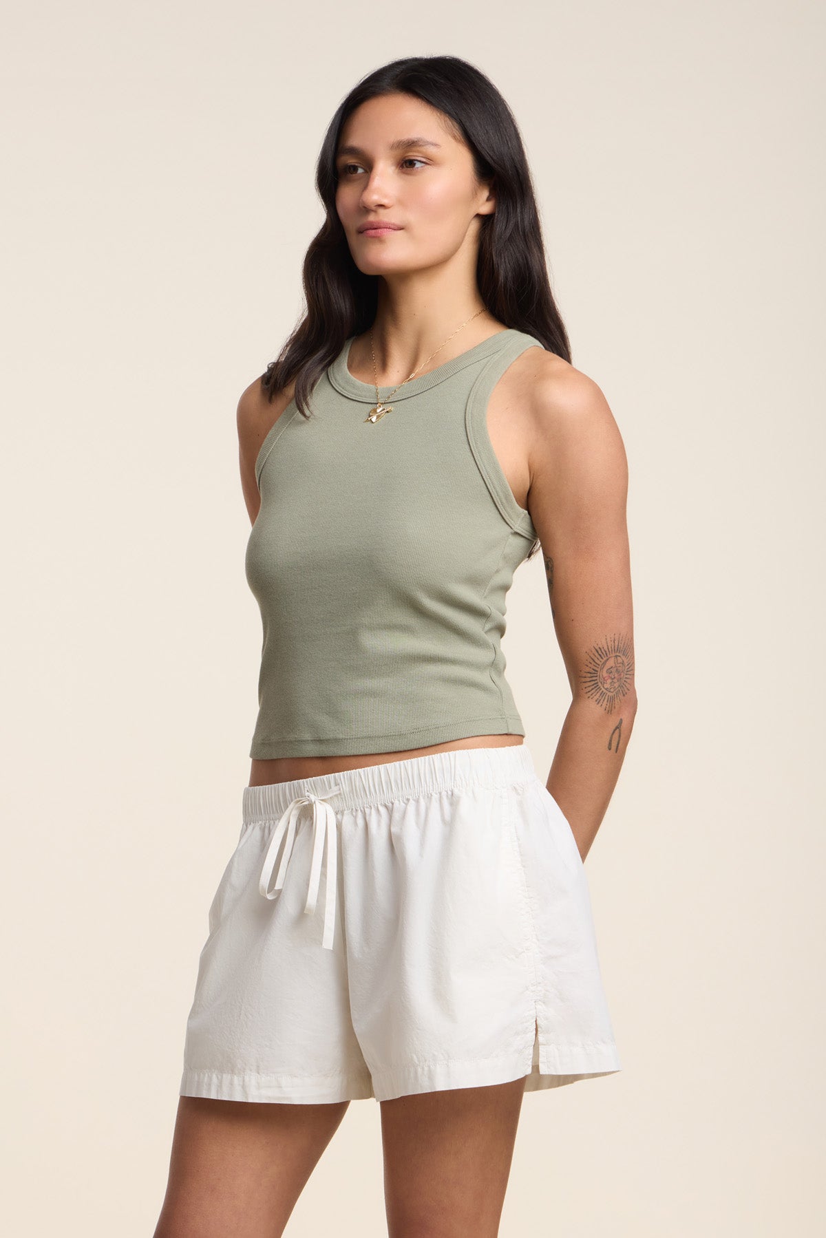 A woman stands against a plain background in the SARI TANK TOP by Velvet by Graham & Spencer, a light green ribbed cotton cropped tank paired with white drawstring shorts—a modern essential look, showcasing a left arm tattoo.