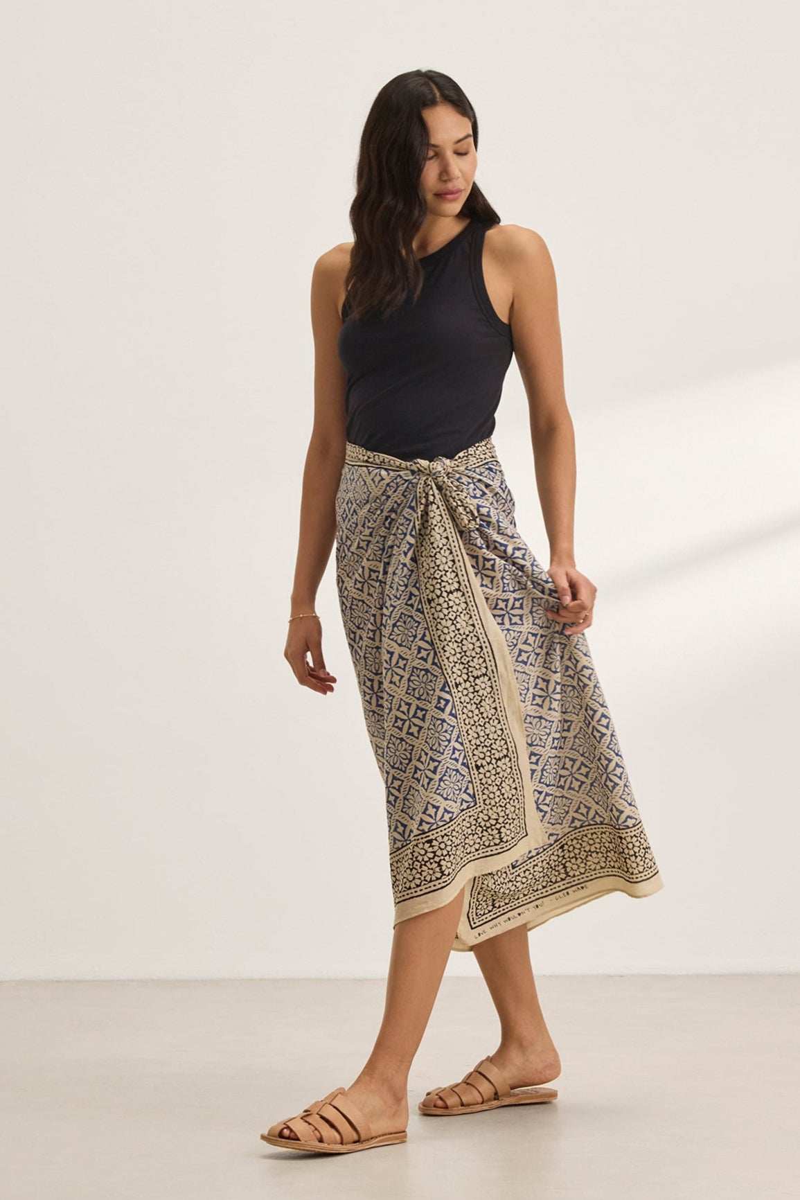   A person stands wearing a black sleeveless top, the JENNA SARONG by Velvet by Graham & Spencer, and sandals, effortlessly combining style and simplicity against a plain background. 