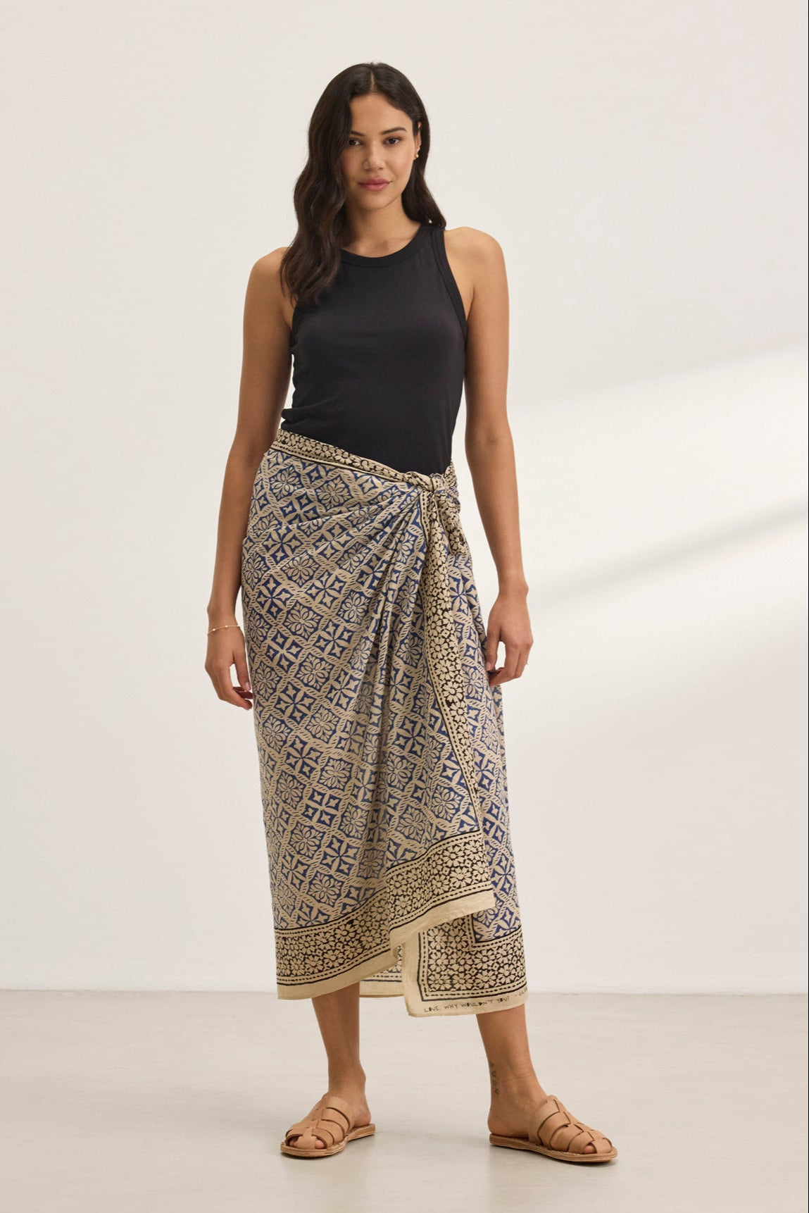   A woman wearing Velvet by Graham & Spencer's dark sleeveless top and the vibrant print JENNA SARONG stands on a light-colored floor in sandals, set against a plain white background. 