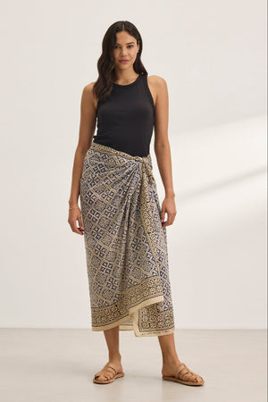 A woman wearing Velvet by Graham & Spencer's dark sleeveless top and the vibrant print JENNA SARONG stands on a light-colored floor in sandals, set against a plain white background.