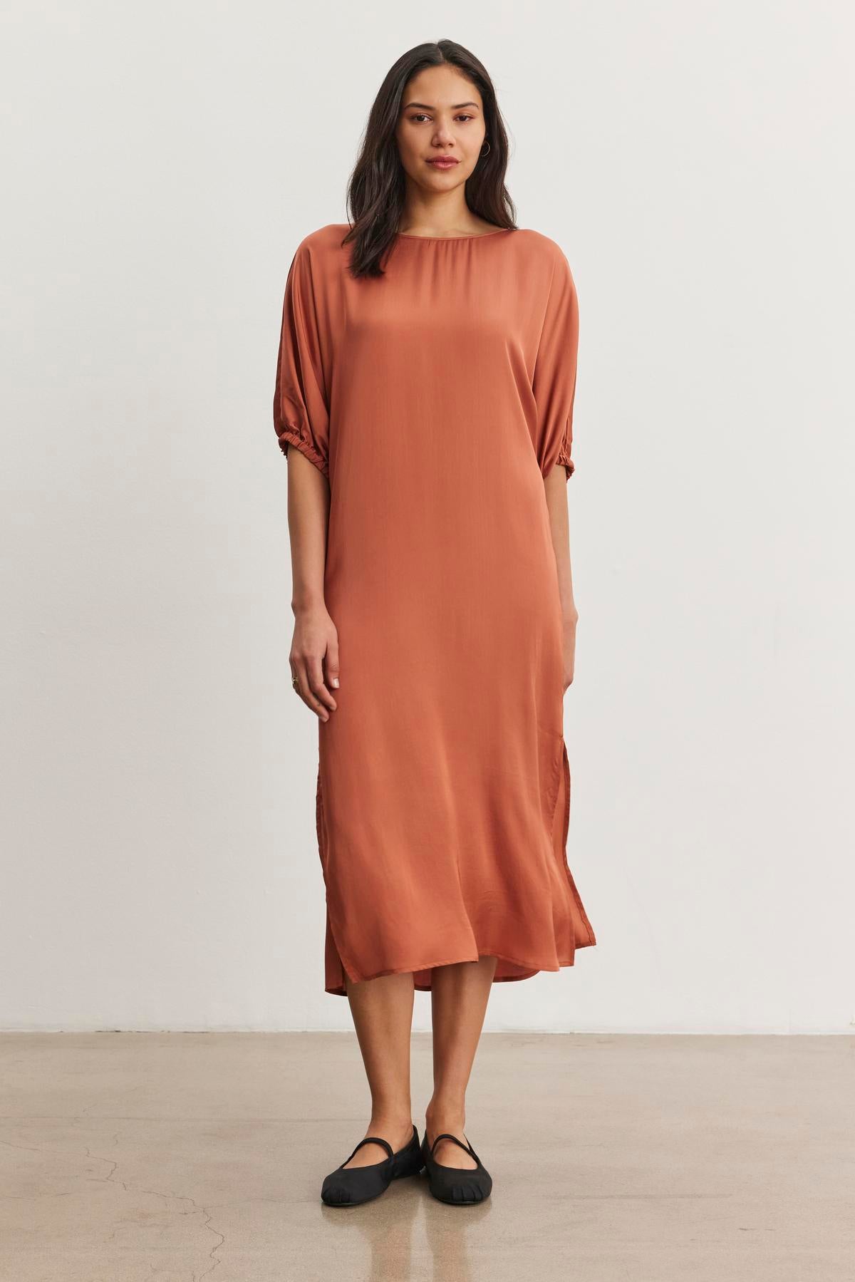   A woman stands against a plain background wearing the LAUREL SATIN DRESS by Velvet by Graham & Spencer, featuring a rust color and dolman sleeves, paired with black shoes. 