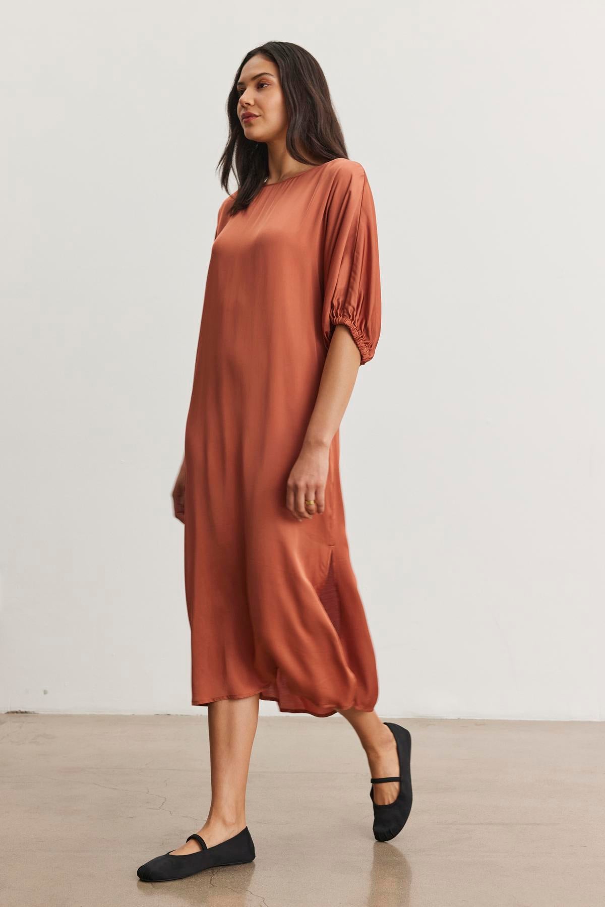   A woman in the LAUREL SATIN DRESS by Velvet by Graham & Spencer, styled with dolman sleeves and rust color, pairs the look with black flats as she walks through a minimalist setting. 