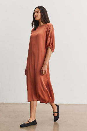 A woman in the LAUREL SATIN DRESS by Velvet by Graham & Spencer, styled with dolman sleeves and rust color, pairs the look with black flats as she walks through a minimalist setting.