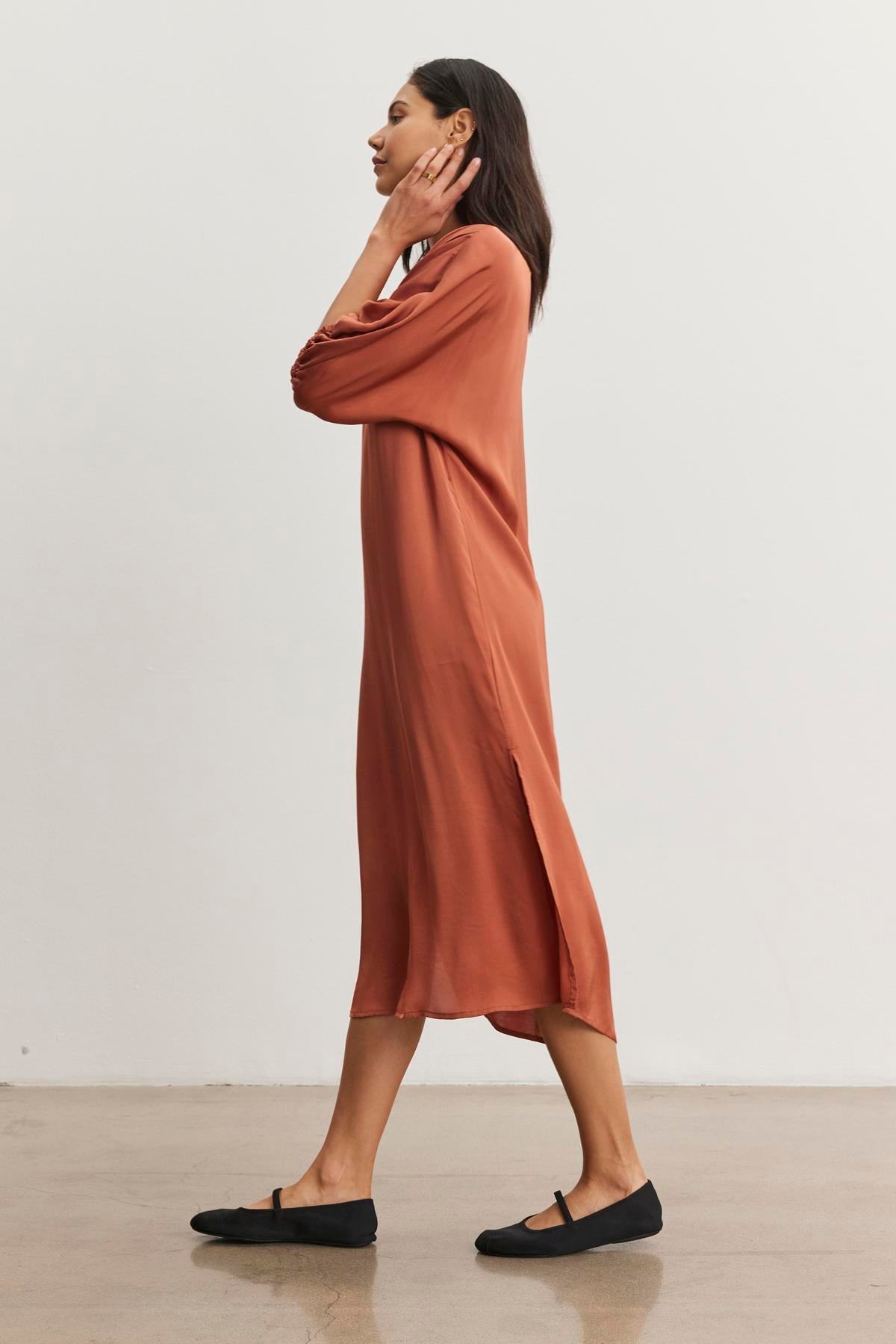   Wearing the LAUREL SATIN DRESS by Velvet by Graham & Spencer, a person stands sideways against a plain background. The dress, characterized by its rust color and dolman sleeves, pairs perfectly with black flats as they touch their face with one hand. 