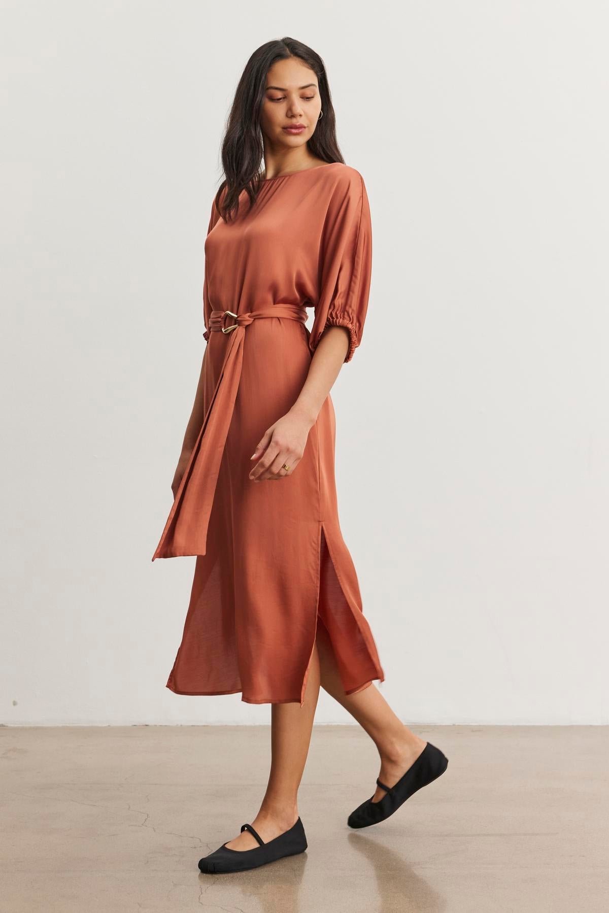   A woman gracefully walks on a smooth floor, wearing the rust-colored LAUREL SATIN DRESS by Velvet by Graham & Spencer, featuring dolman sleeves and a stylish detachable belt. Her look is completed with classic black flats. 