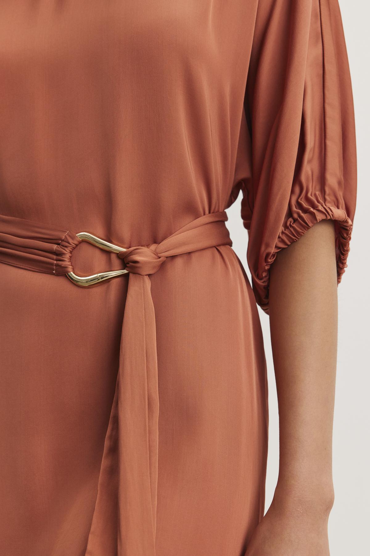   The LAUREL SATIN DRESS from Velvet by Graham & Spencer features a rust-colored satin viscose fabric with dolman sleeves, complemented by a matching detachable belt with a gold buckle. 