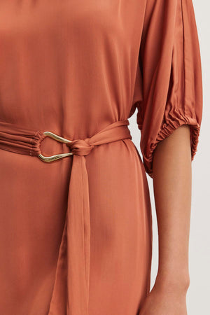 The LAUREL SATIN DRESS from Velvet by Graham & Spencer features a rust-colored satin viscose fabric with dolman sleeves, complemented by a matching detachable belt with a gold buckle.