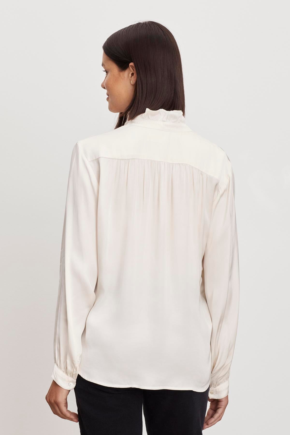   A woman wearing a cream ALI BUTTON FRONT BLOUSE with a ruffle neckline is seen from the back by Velvet by Graham & Spencer. 