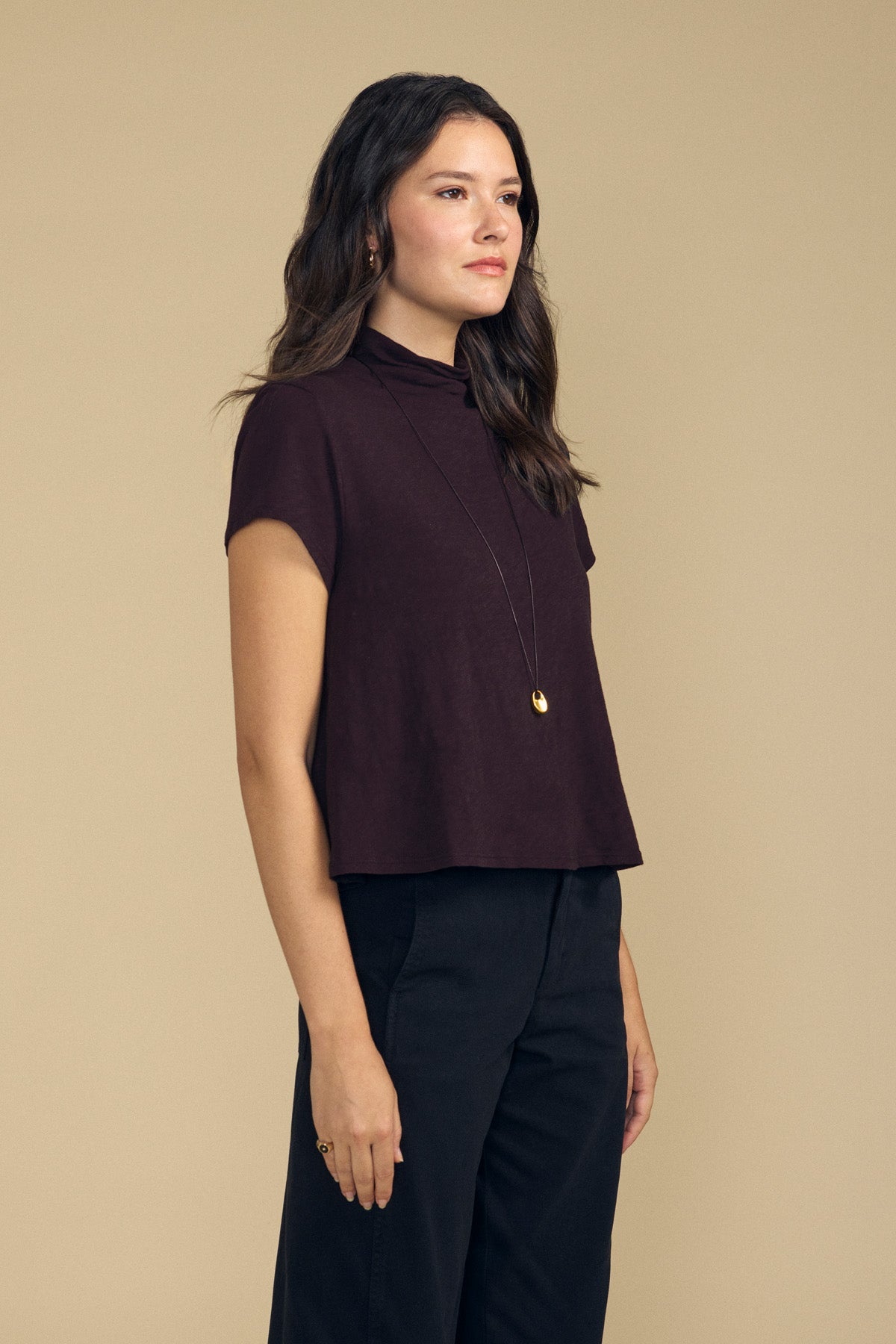 A person wearing a dark brown, boxy silhouette Velvet by Graham & Spencer SAVI TEE with a mock neck, paired with a long gold pendant necklace and black trousers.-46461710958785
