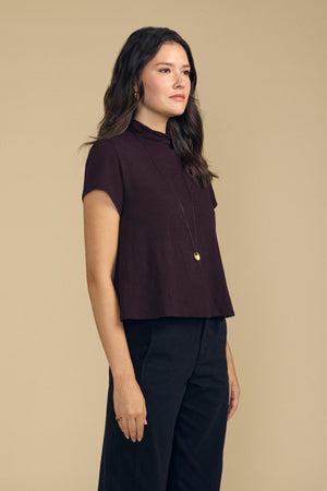 A person wearing a dark brown, boxy silhouette Velvet by Graham & Spencer SAVI TEE with a mock neck, paired with a long gold pendant necklace and black trousers.