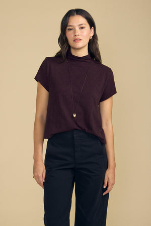 A person wearing a dark purple cotton slub SAVI TEE by Velvet by Graham & Spencer with a relaxed turtleneck, paired with black trousers and a long pendant necklace.