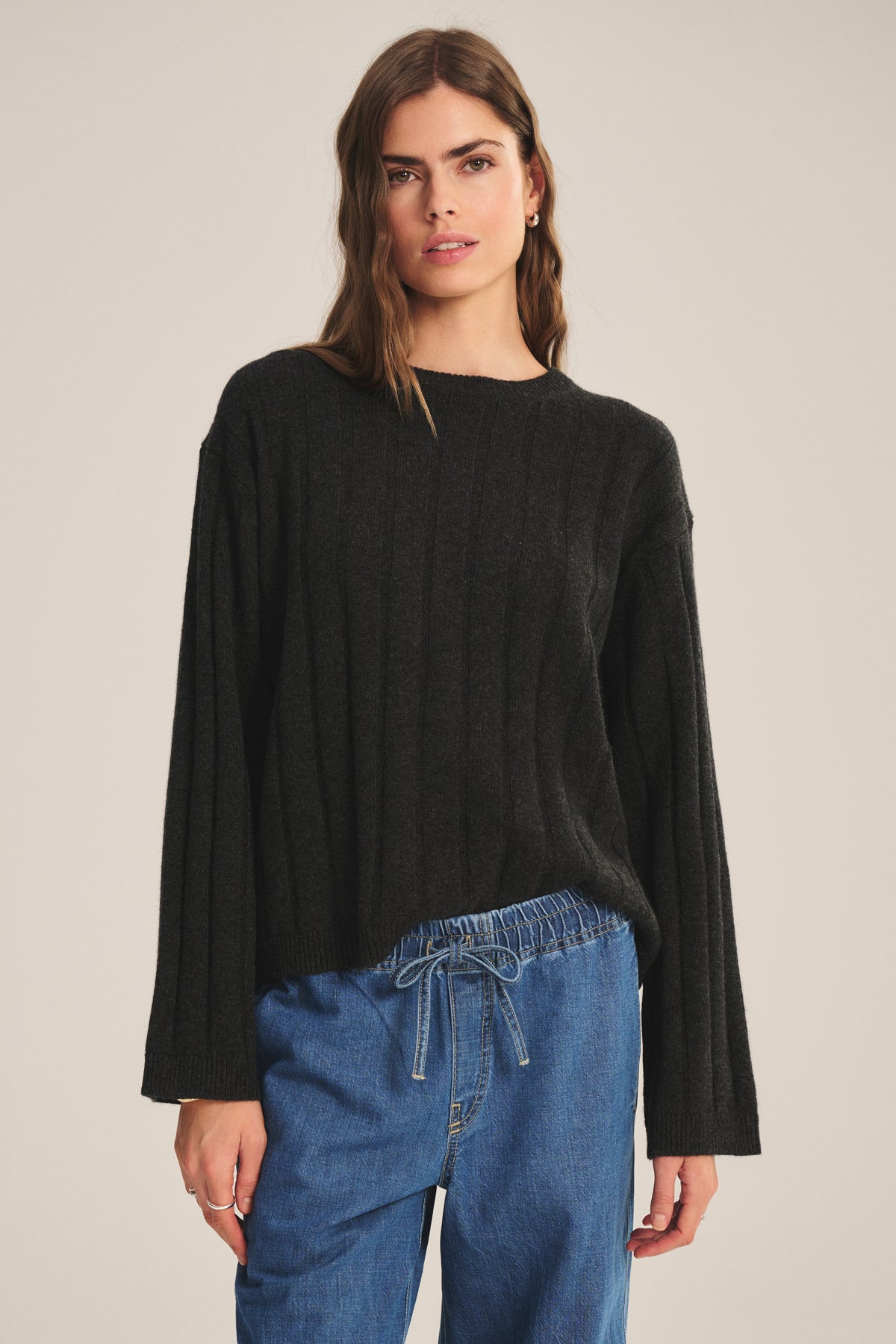 A person stands against a plain background wearing the SAWTELLE CASHMERE SWEATER by Velvet by Jenny Graham, a loose-fitting black ribbed sweater, paired with blue drawstring denim pants.-39838371872961